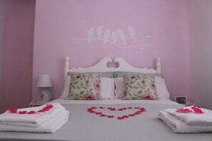 Een slaapkamer met een bed, kussens, een nachtkastje, een lamp en een hartvormige decoratie op het bed.