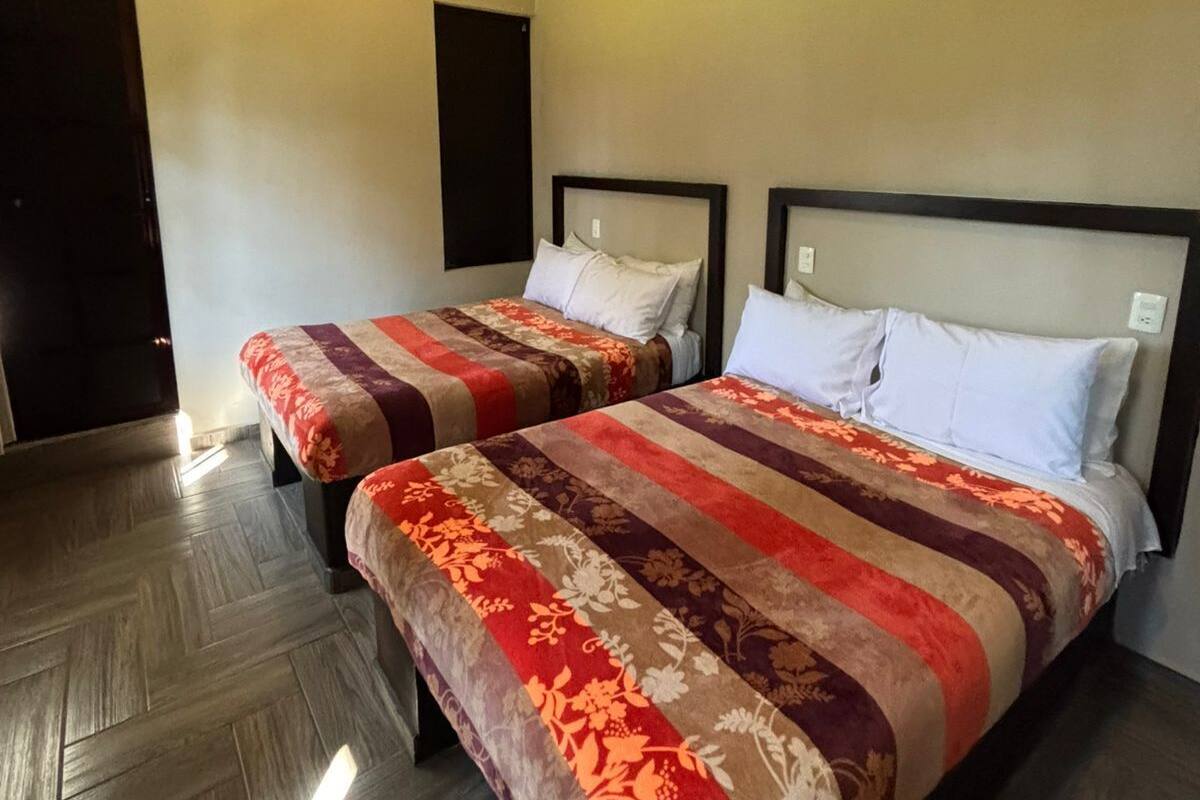 A hotel room with two beds, a wooden floor, a ceiling light, and an air conditioning unit.