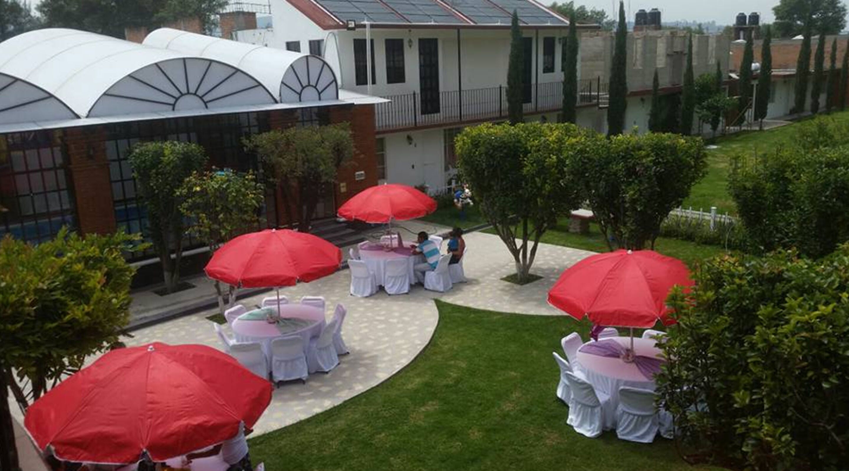 An outdoor patio area with round tables and red umbrellas, surrounded by greenery and a building with a modern design.