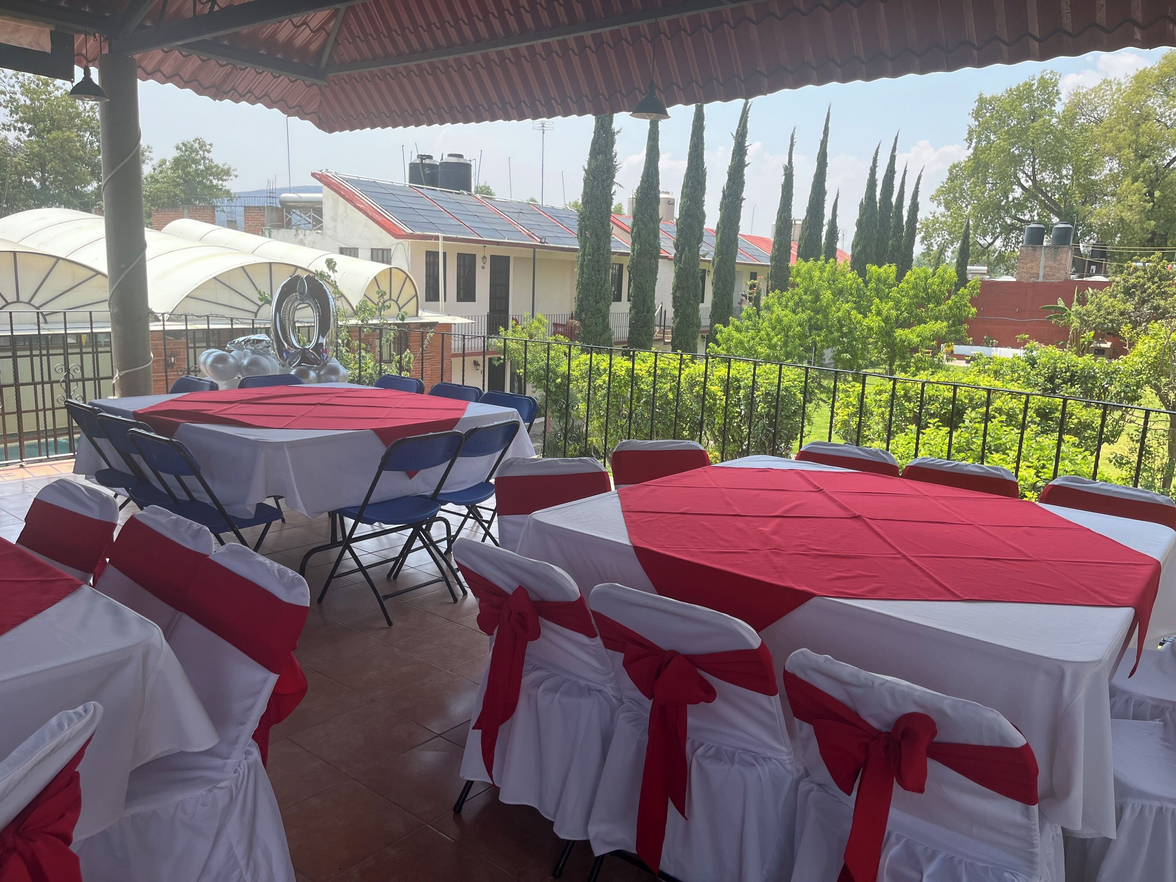 An outdoor patio with round tables covered in white and red tablecloths, chairs with white and red sashes, and a view of buildings and trees in the background.