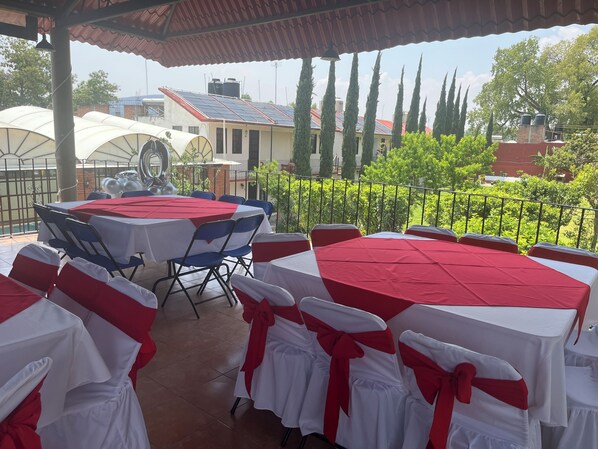 Un patio al aire libre con mesas redondas cubiertas con manteles blancos y rojos, sillas con cintas blancas y rojas, y una vista de edificios y árboles al fondo.