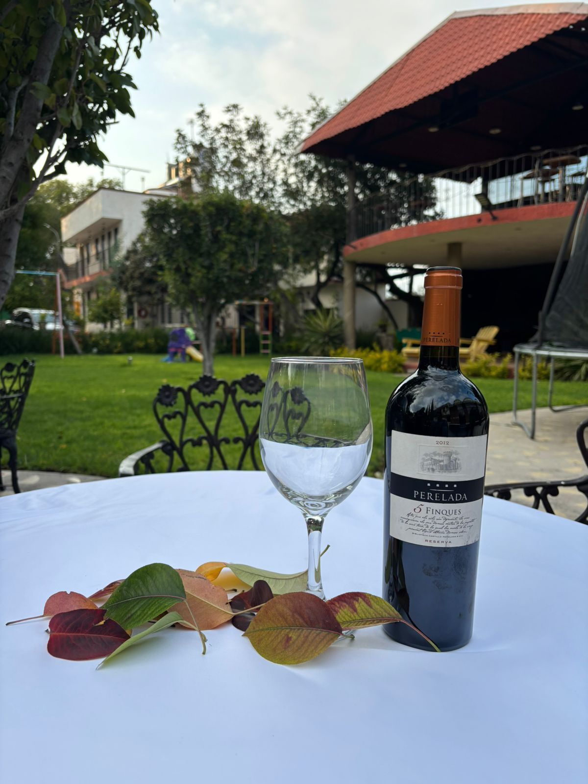 A bottle of wine and a glass on a table outdoors with a building and greenery in the background.