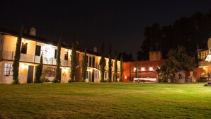 A two-story building with a well-lit exterior, a grassy lawn in the foreground, and a brick wall with a window on the right side. - Hotel Real Campestre Tepetoci (Tepeji del Río de Ocampo)