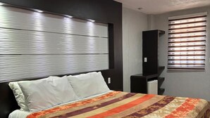 A modern bedroom with a large bed, a headboard with horizontal lines, a bedside table, and a window with blinds. - Hotel Real Campestre Tepetoci (Tepeji del Río de Ocampo)