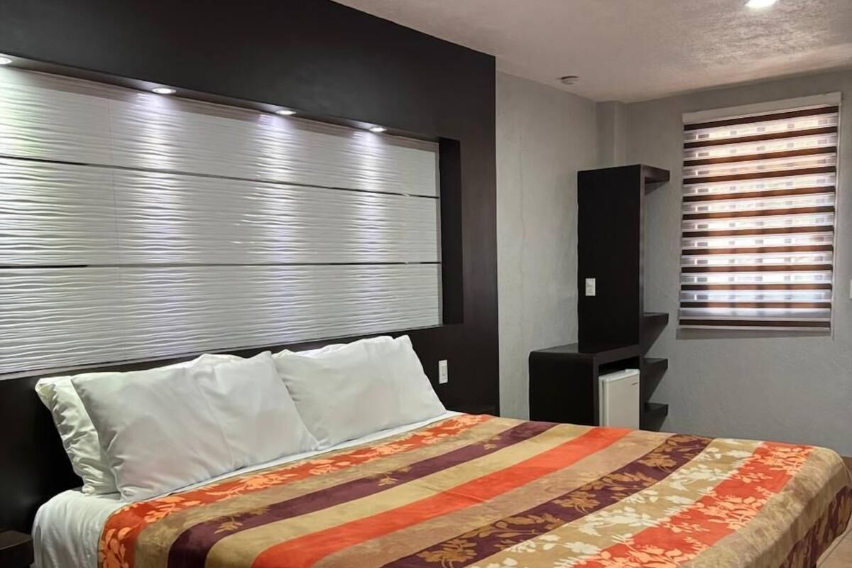 A modern bedroom with a large bed, a headboard with horizontal lines, a bedside table, and a window with blinds.