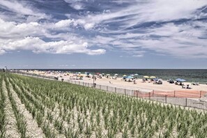 Ein Strand mit Grasreihen im Vordergrund, ein Sandstrand mit Sonnenschirmen und Menschen sowie ein Zaun, der Strand und Gras trennt.
