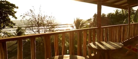 Una terraza de madera con dos mesas redondas y sillas, con vista a una playa y el océano.
