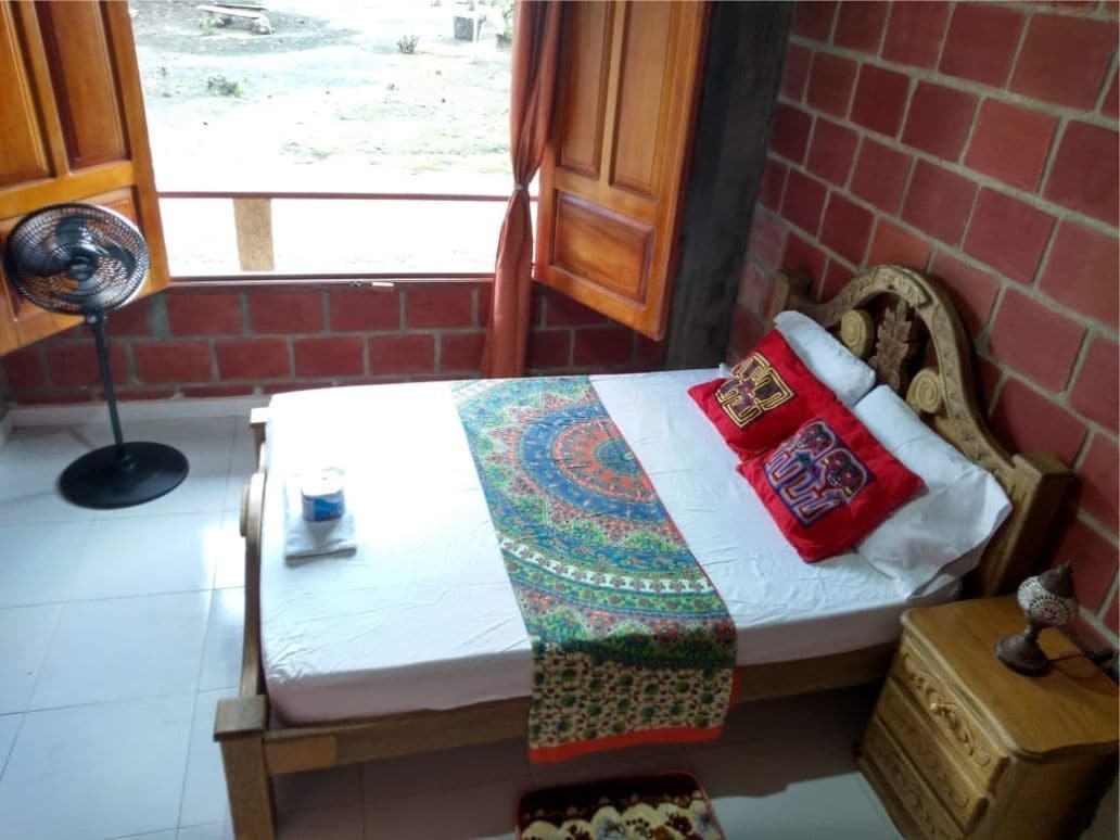 Een kamer met een bed, een houten nachtkastje, een kleine tafel, een ventilator en een raam met gordijnen.