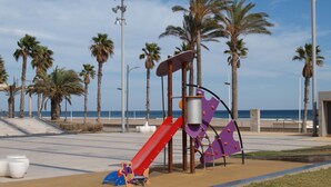 A playground with a red slide and purple climbing structure, surrounded by palm trees and a beach in the background. - ApartUP Patacona Beach Select (Alboraya)