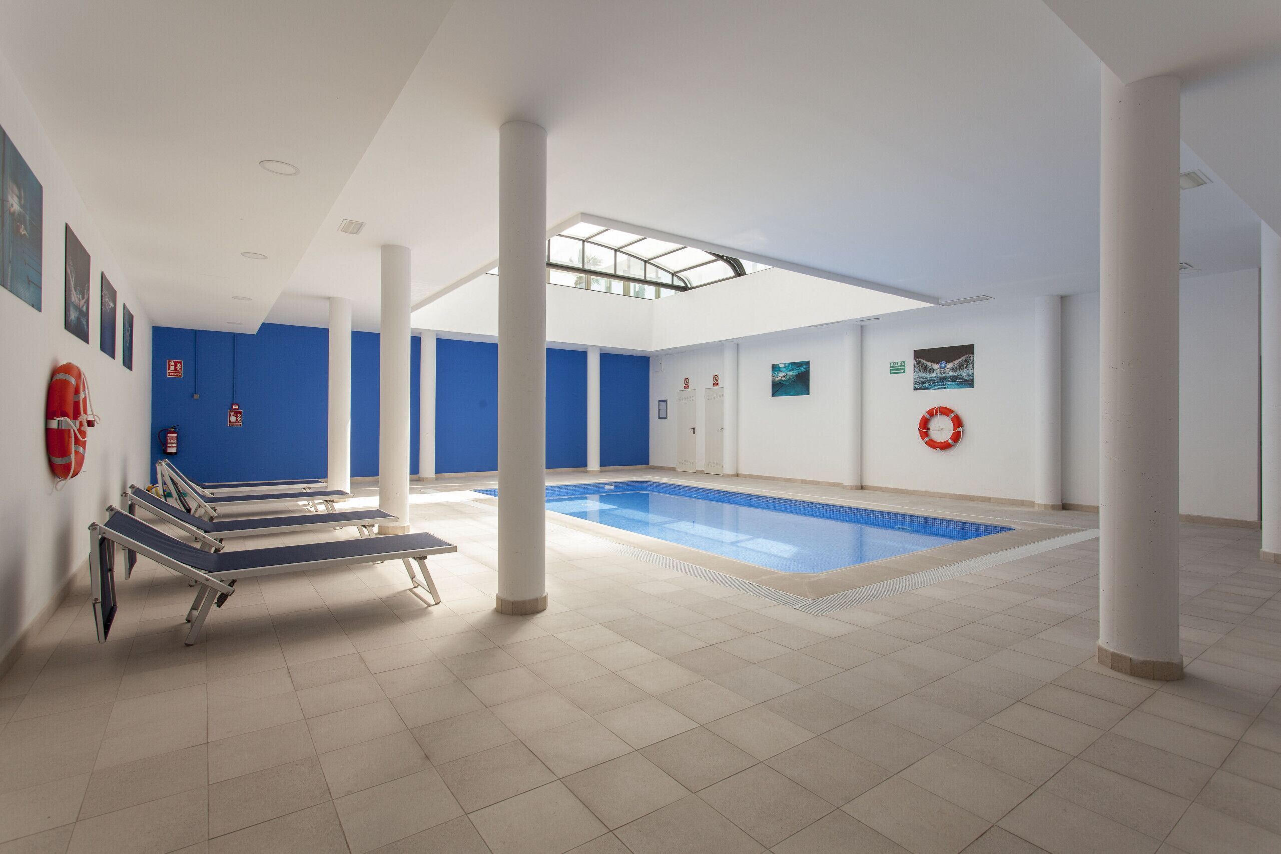 A large indoor swimming pool with clear blue water, surrounded by white pillars and blue walls.