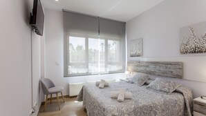 A modern bedroom with a large bed, a chair, a TV mounted on the wall, and a window with blinds. - ApartUP Patacona Beach Select (Alboraya)