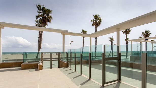 A modern outdoor terrace with glass railings, palm trees, and a view of the beach. - ApartUP Patacona Beach Select (Alboraya)