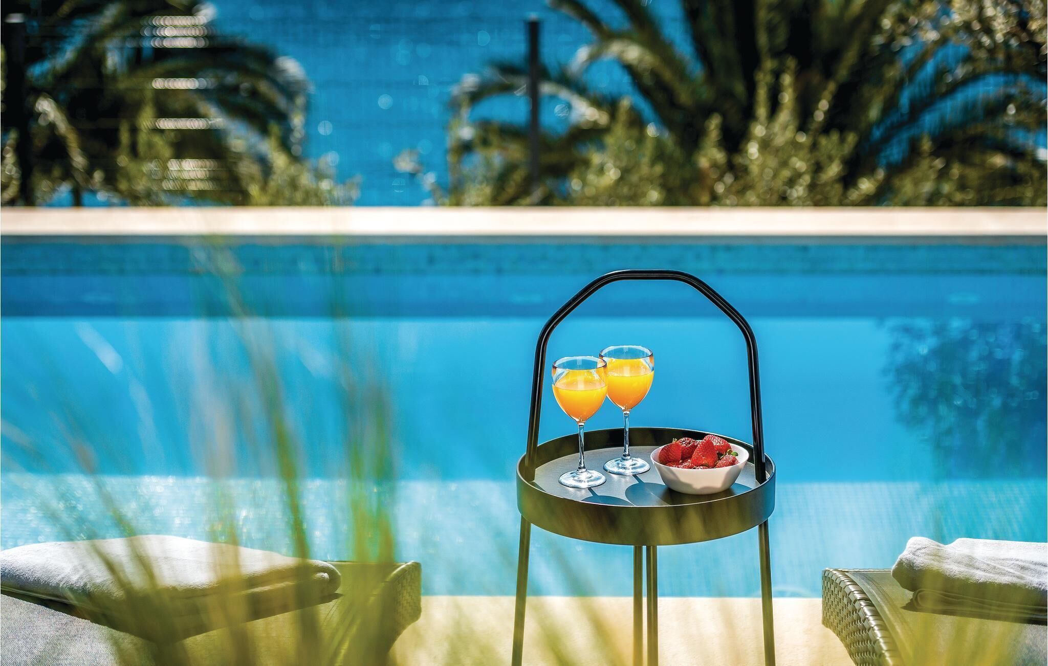 A poolside table with two glasses of orange juice and a bowl of strawberries.