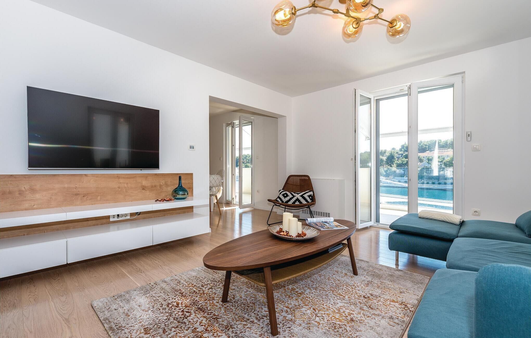 A modern living room with a flat-screen TV, wooden console, a round coffee table, a blue sofa, and a view of a body of water through large sliding doors.