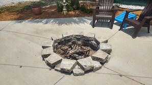 Un hoyo para fuego de hormigón con un borde de piedra, rodeado por un patio con sillas y un banco azul.