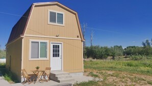 Une petite maison de plain-pied, à l’extérieur de couleur beige et au toit rouge. Elle possède une porte d’entrée avec un panneau en verre et une fenêtre avec des volets. On peut voir une petite table et deux chaises devant la maison.