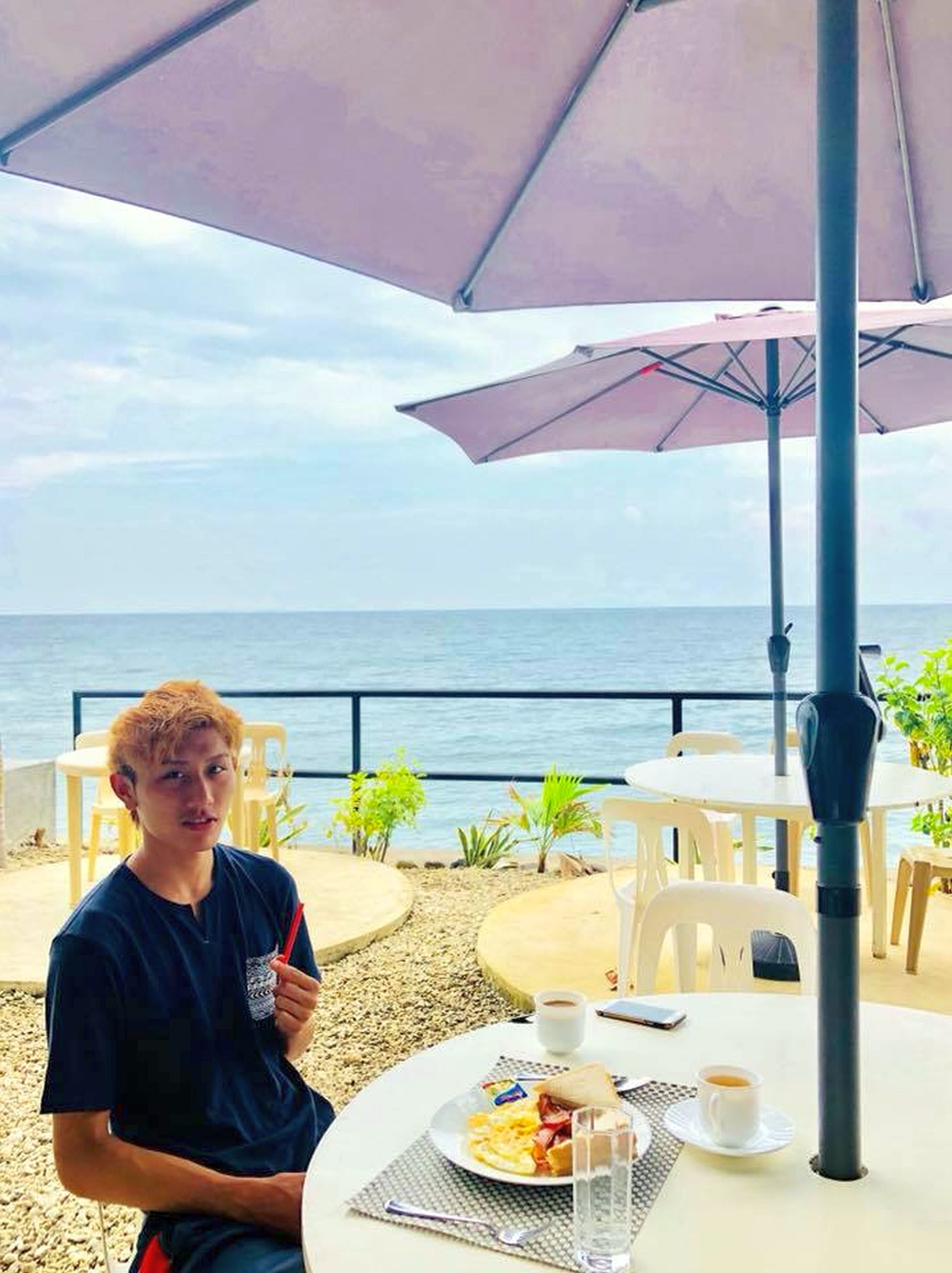 Une personne assise à une table en plein air, avec vue sur la mer, devant un repas et une boisson.