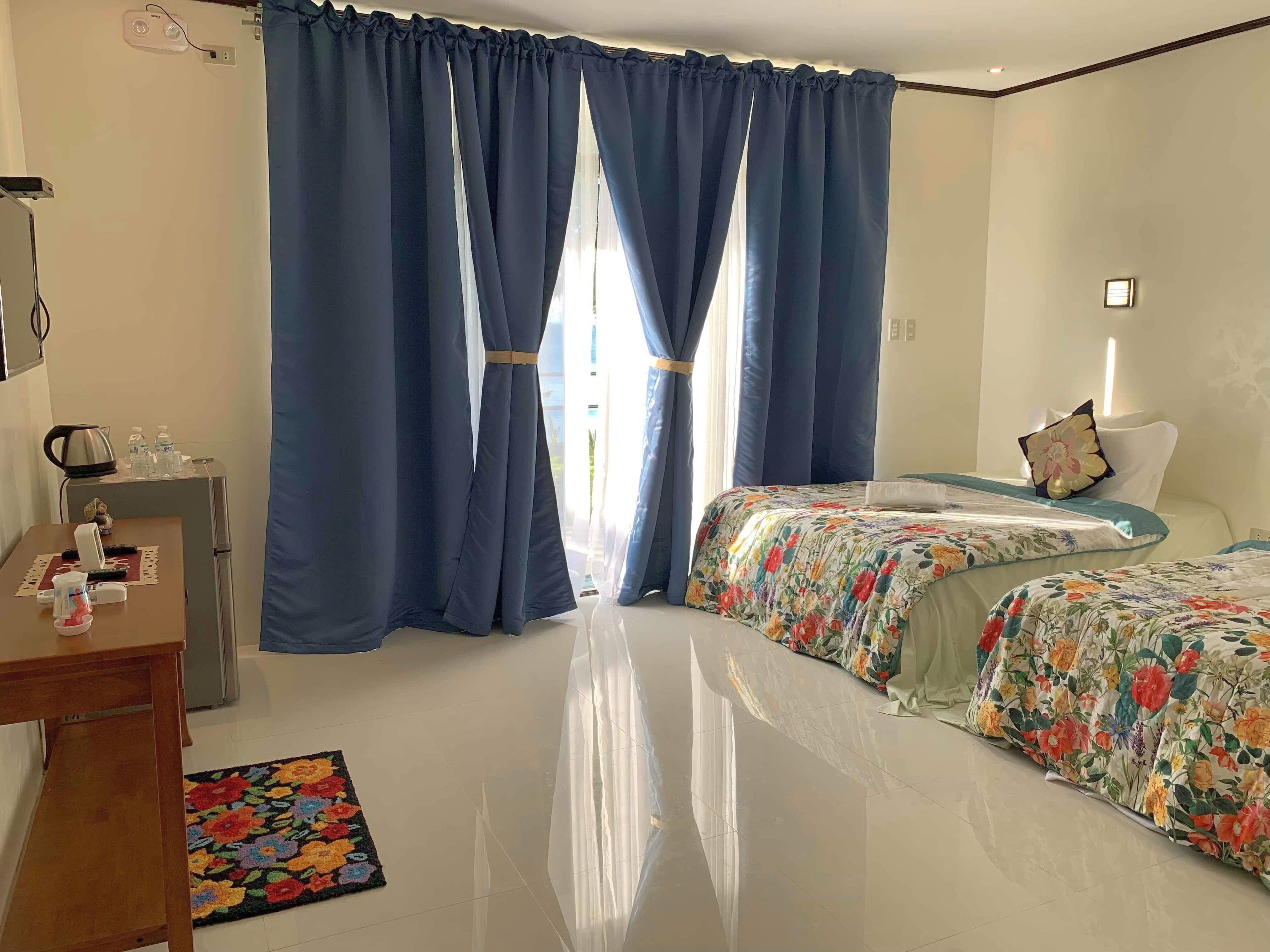A hotel room with two beds, a desk, a small kitchenette, and a large window with blue curtains.