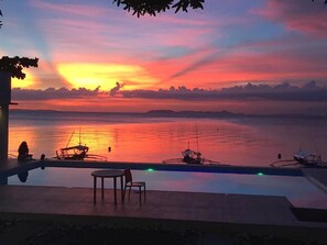 Vista al atardecer desde una piscina infinita con vista a un mar tranquilo, barcos y una costa lejana.