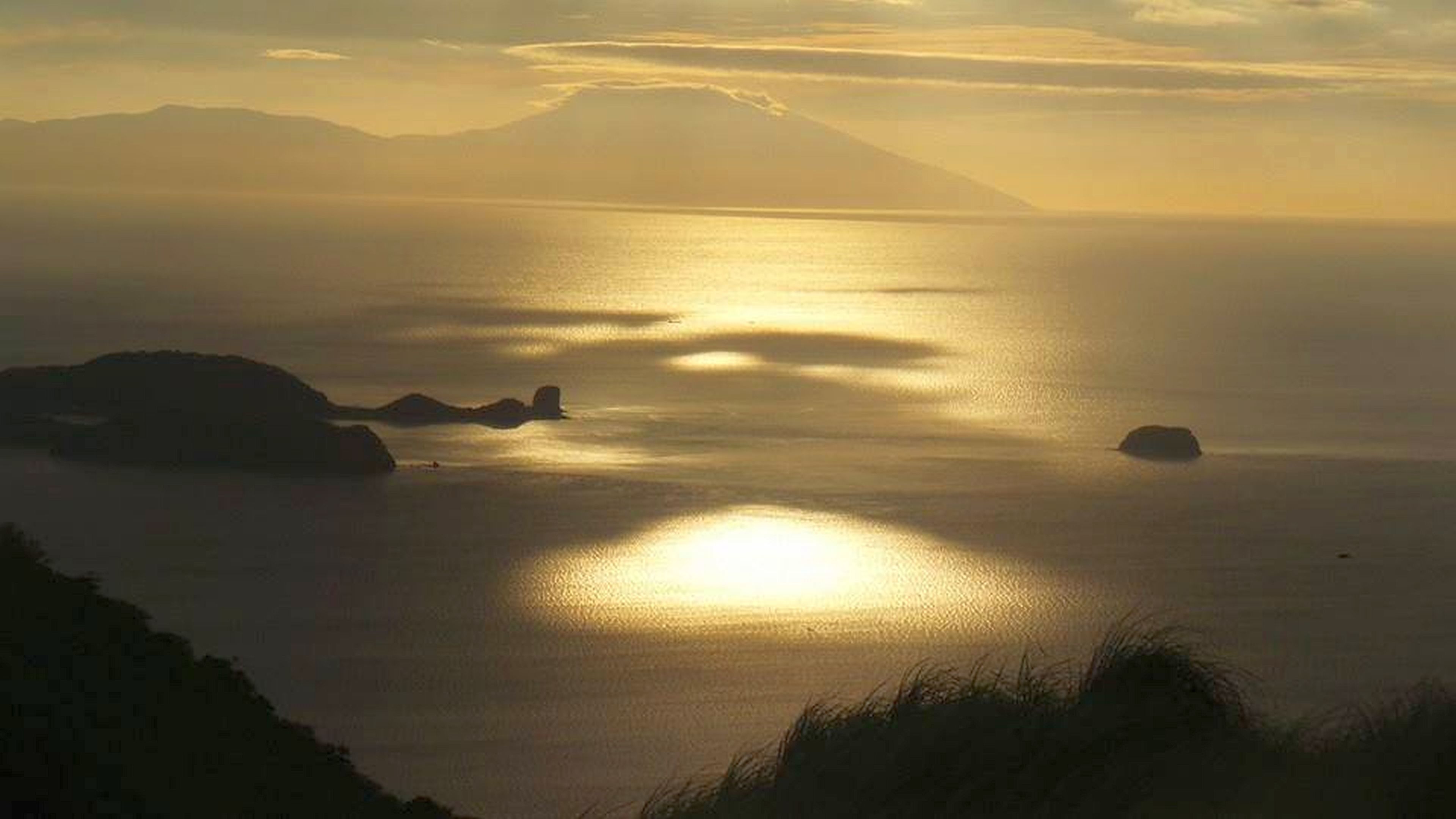 Paisaje costero con aguas tranquilas reflejando la luz del sol, montañas lejanas y pequeñas islas.