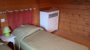 Une petite pièce de cabane en bois comprenant deux lits simples, une table de chevet avec une lampe et une commode blanche.