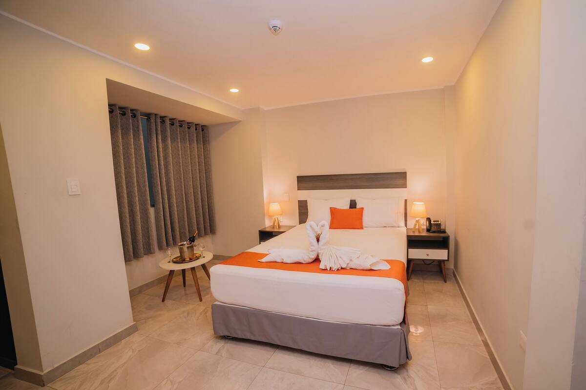 Une chambre d’hôtel avec un grand lit, deux tables de chevet, une petite table, une lampe et un rideau.