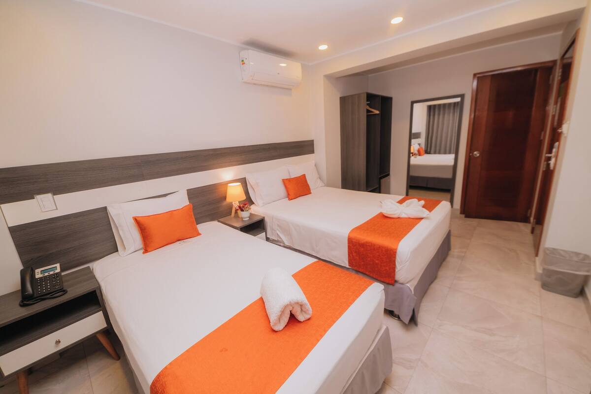 Une chambre d’hôtel avec deux lits, chacun recouvert d’une couvre-lit orange et doté d’oreillers blancs. Il y a une table de chevet avec une lampe et un téléphone. La chambre est équipée d’une tête de lit en bois, d’un miroir et d’une porte donnant sur une autre pièce.