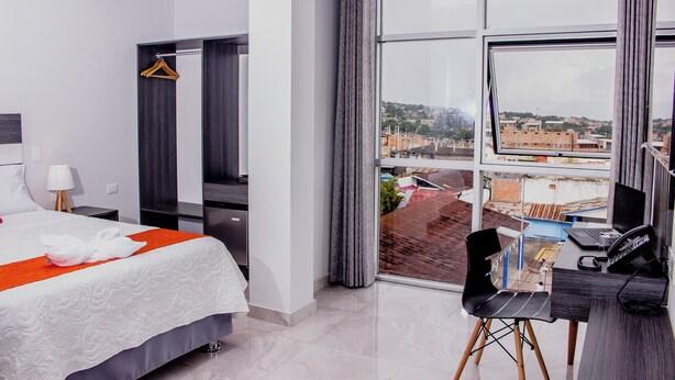 A hotel room with a bed, a desk with a computer, a chair, and a view of the city through large windows. - Palm Hotel (Tarapoto)