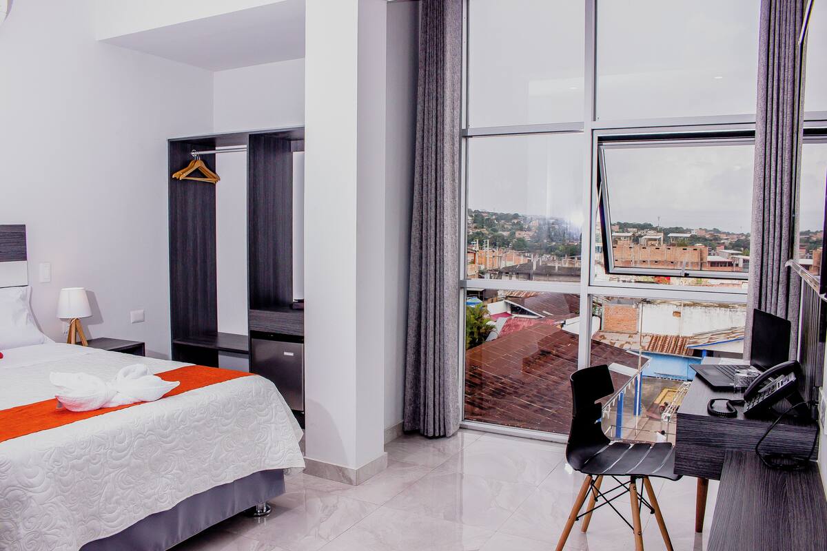 Une chambre d’hôtel avec un lit, un bureau équipé d’un ordinateur, une chaise et une vue sur la ville grâce à de grandes fenêtres.