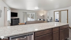 A modern kitchen with granite countertops, a dishwasher, and wooden cabinets. There is a large window with curtains, a sofa, and a clock on the wall. - Matanuska Villa #1, Glacier Suites - Matanuska Villa #1 (Matanuska-Susitna)