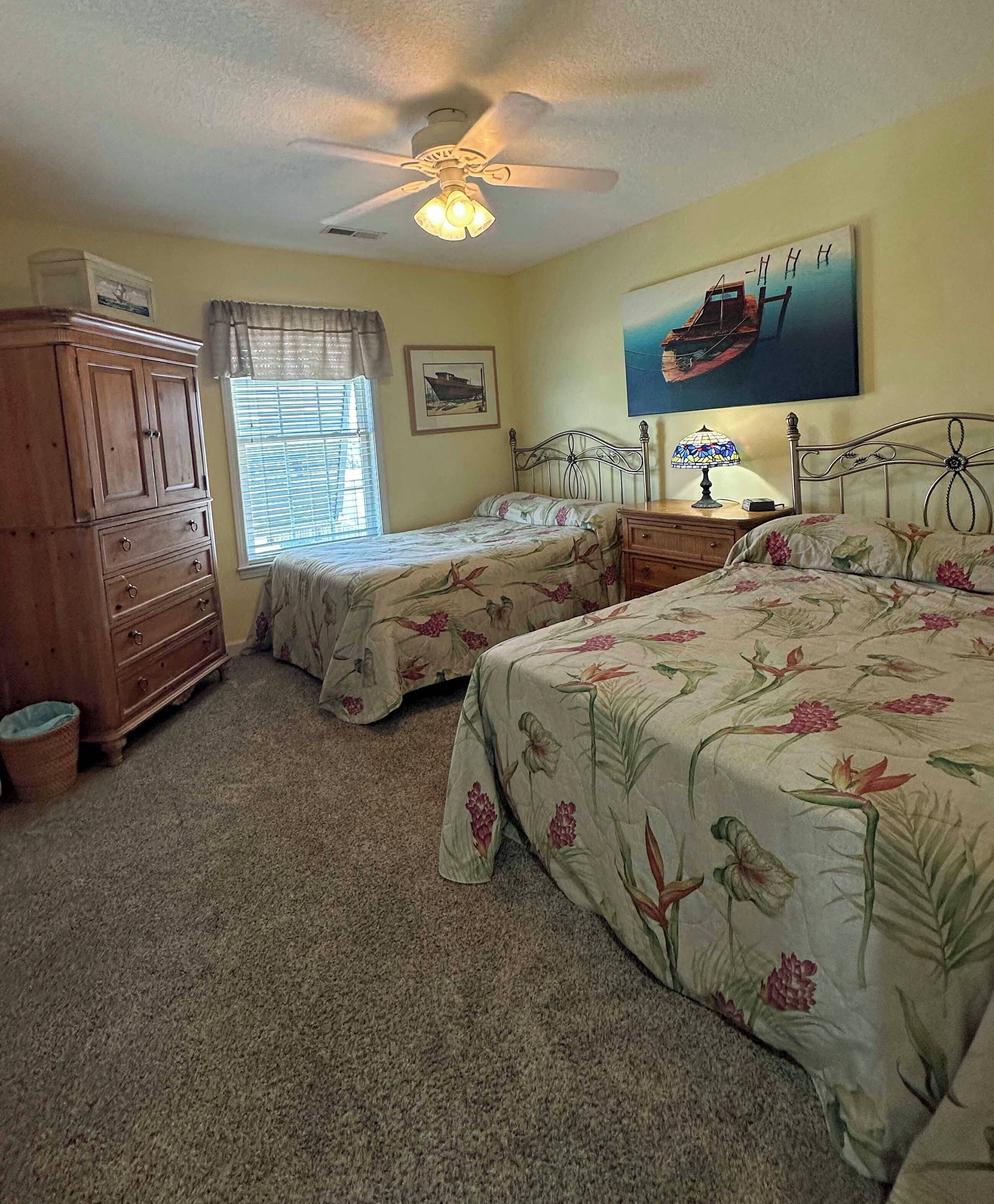 Une chambre avec deux lits simples, une commode en bois, un ventilateur de plafond et un tableau représentant un bateau accroché au mur.