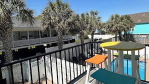 Eine Holzterrasse mit Pool, weißem Zaun und Blick auf ein Gebäude mit Balkon.