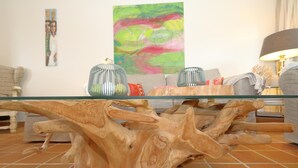 Uma sala de estar com uma mesa de centro de vidro apoiada em uma base de madeira com design escultural, um quadro abstrato e colorido na parede e uma luminária sobre uma mesa lateral.