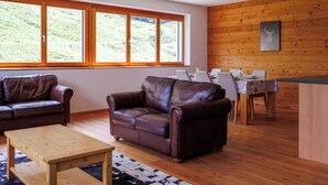 Ein modernes Wohnzimmer mit Ledersofas, einem Holztisch und einem Essbereich mit Tisch für vier Personen. Große Fenster bieten Blick auf eine grüne Landschaft.