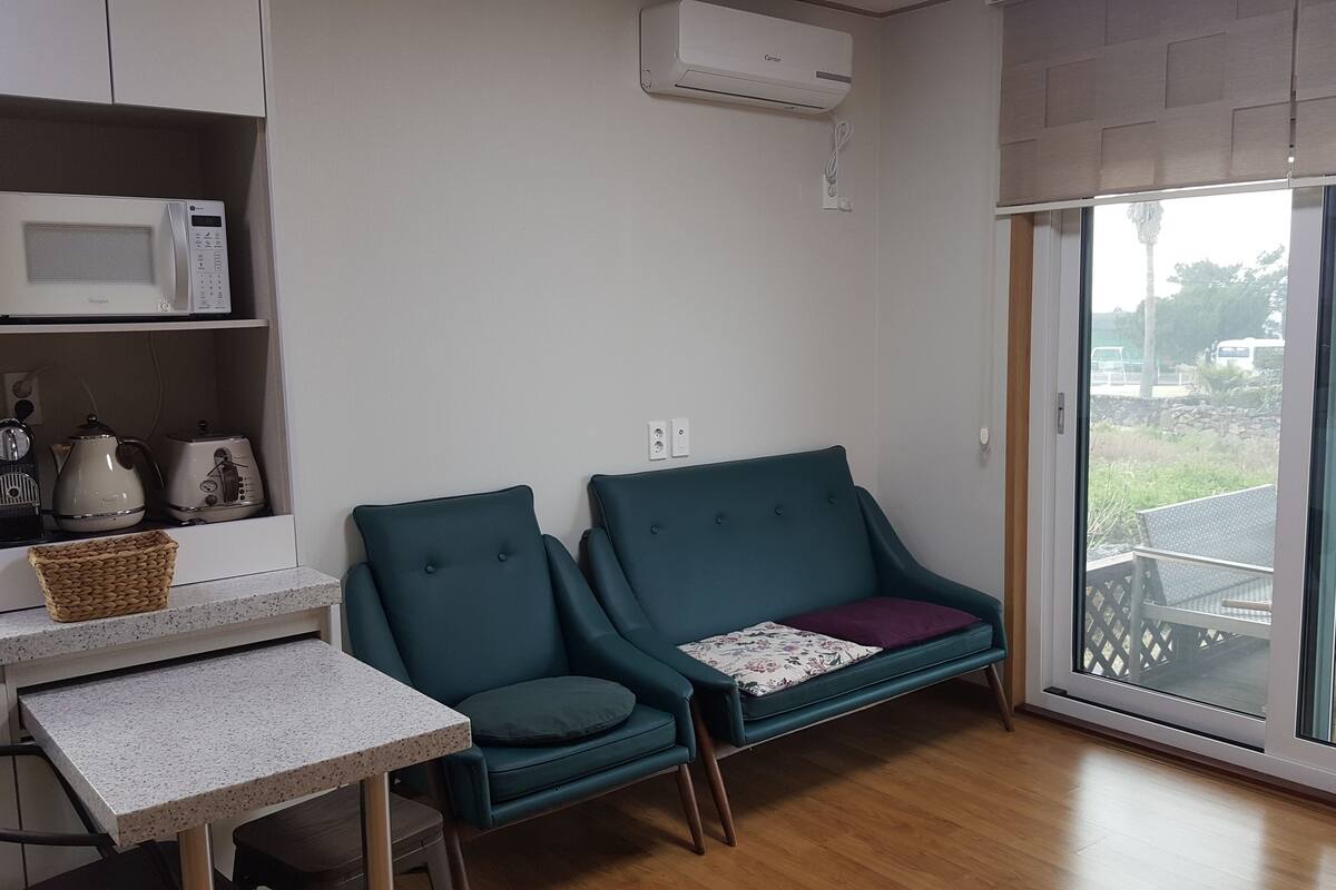 A compact living space with a kitchenette, a teal sofa, a small table, and a sliding glass door leading to an outdoor area.