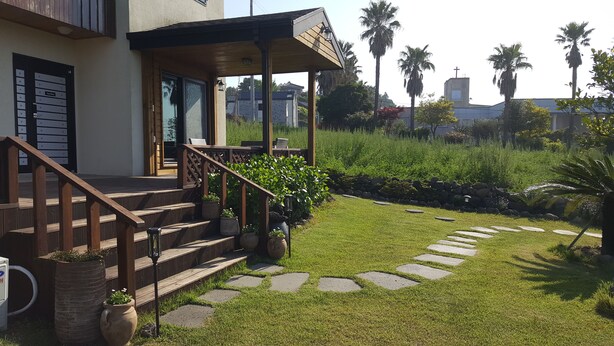 A house with a wooden staircase leading to a covered porch, a stone pathway, and a lush garden with palm trees. - Sonangche (Jeju City)