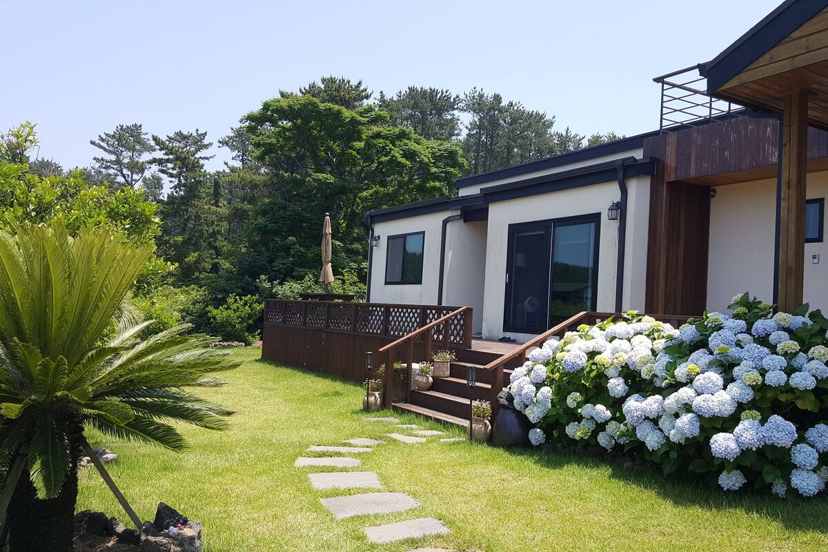 A modern house with a wooden deck, a stone pathway, and a lush garden with hydrangeas.