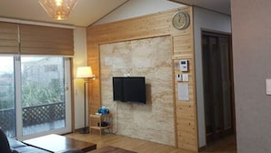 A modern hotel room with a wooden wall, a flat-screen TV, a clock, and a small table with a lamp. - Sonangche (Jeju City)