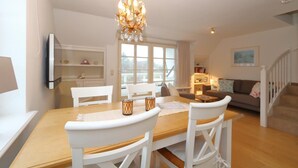 Een eetgedeelte met een houten tafel en witte stoelen, een kroonluchter en uitzicht op een woonkamer met een bank en een trap.