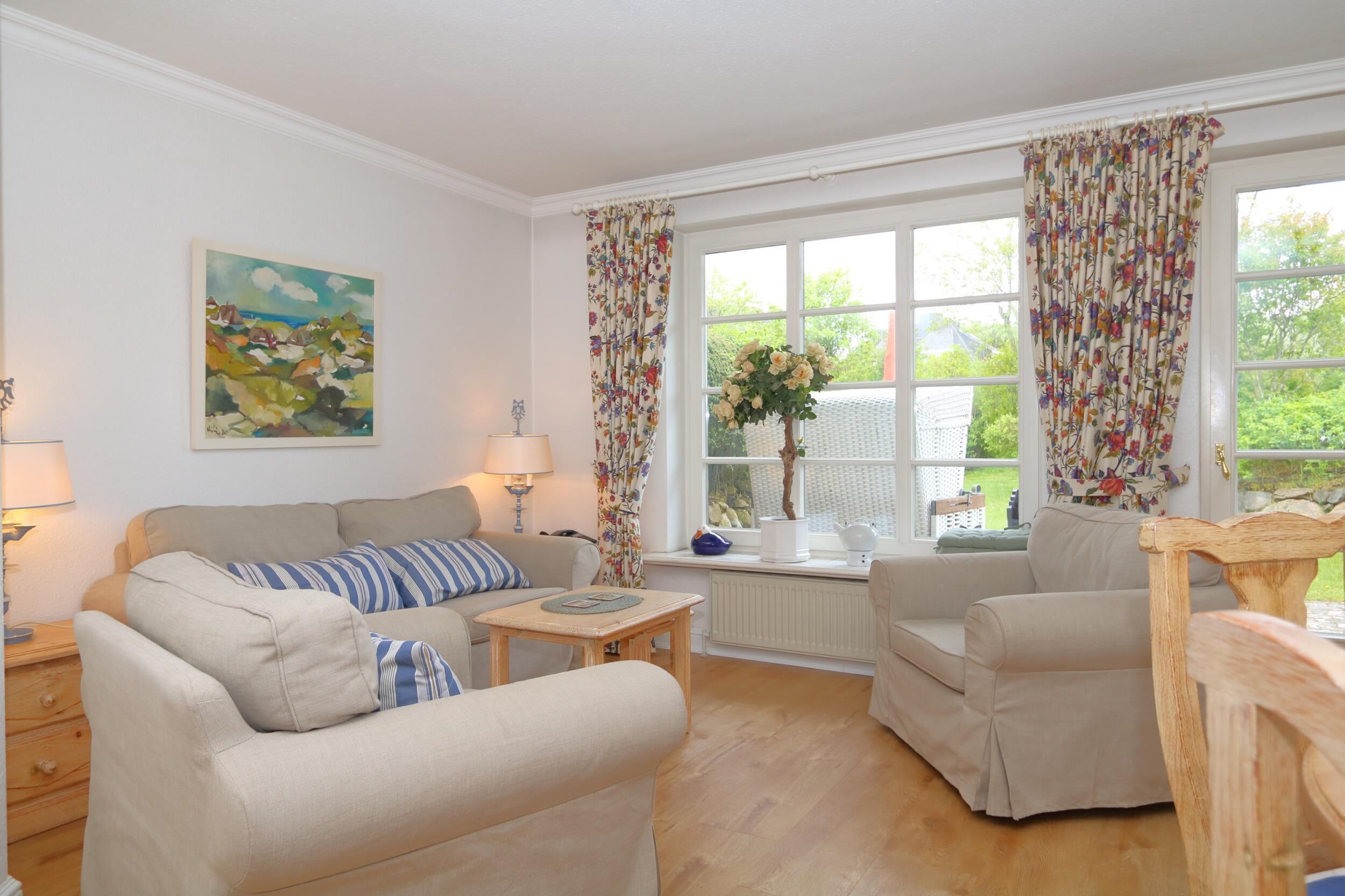 Een gezellige woonkamer met een bank, fauteuil en een kleine tafel. Er is een raam met gordijnen, een schilderij aan de muur en een vaas met bloemen op de vensterbank.