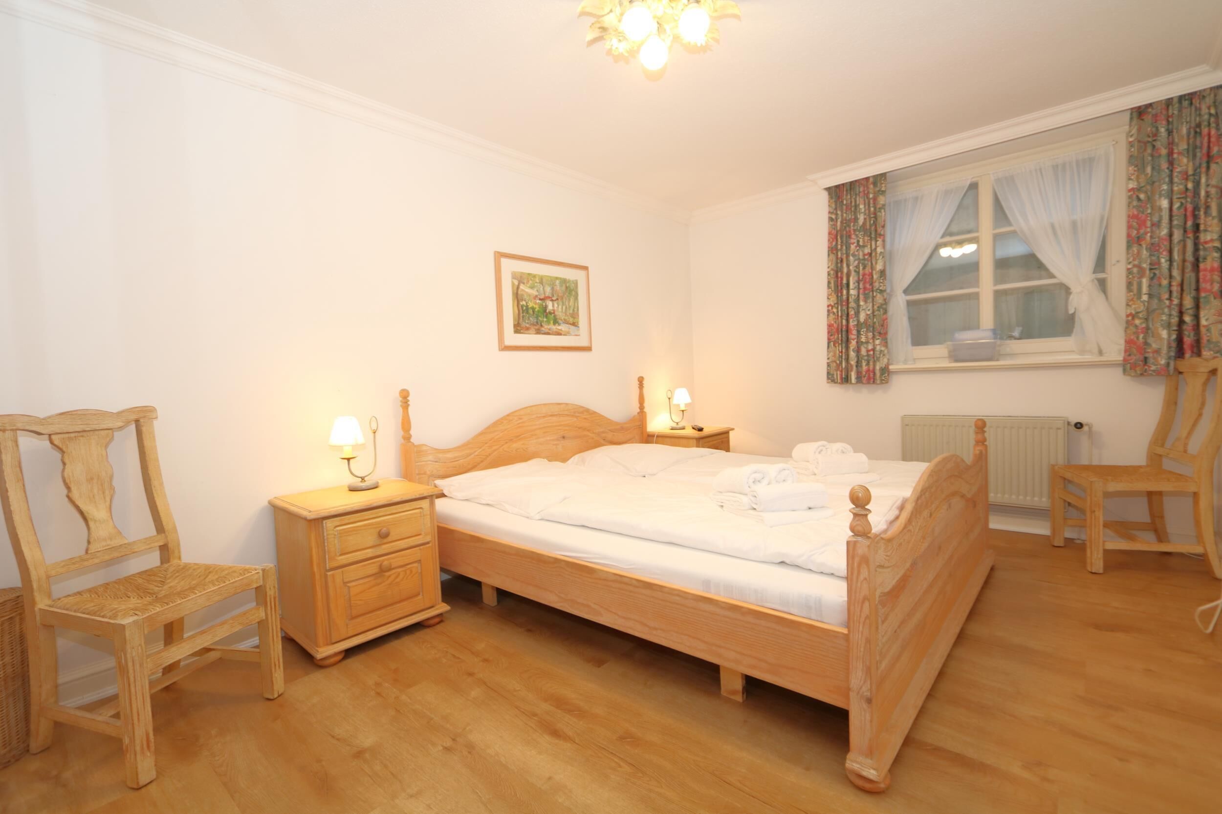 A bedroom with a wooden bed, two bedside tables with lamps, a wooden chair, a radiator, and a window with floral curtains.