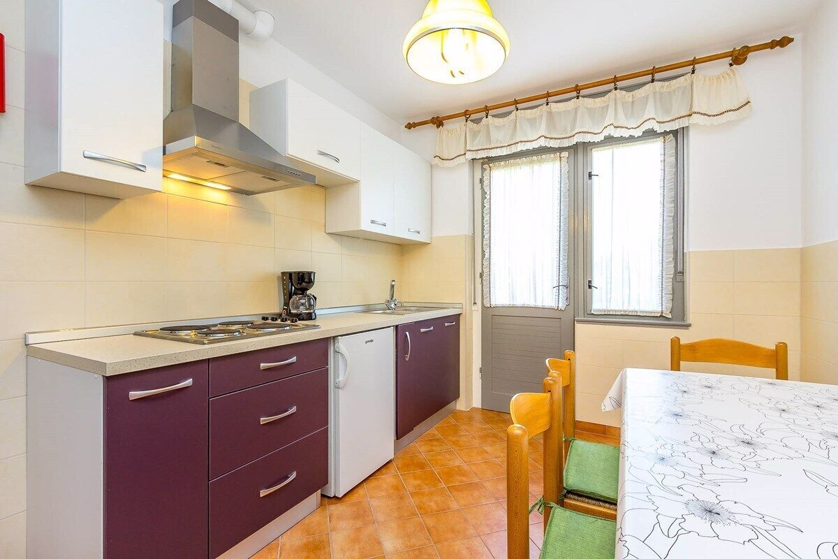 Cocina compacta con estufa, lavaplatos y armarios blancos. Hay una mesa de comedor con sillas y una ventana con cortinas.