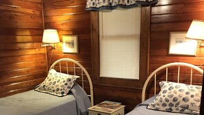 A cozy cabin-style bedroom with two single beds, each with a matching headboard and pillow, a small bedside table with a lamp, and a window with blinds. - Cottage:  3005 Palmetto Blvd. (Edisto Beach)