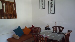 A dining area with a glass-top table, wooden chairs, a brown sofa with colorful cushions, and a small black vase with dried plants. - 1 and 1 (Malimbada)