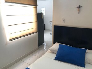 A hotel room with a bed, a nightstand, a wall-mounted cross, and a window with blinds. - Cozy 1 room apartment in Laureles (Medellín)