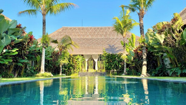 Una piscina cristalina con un patrón geométrico de azulejos, rodeada de exuberante follaje tropical y una estructura con techo de paja.