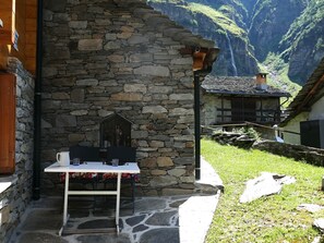 Ein Steinhaus mit Tisch und Stühlen vor der Tür, einer Holztür und Blick auf einen Berg mit einem Wasserfall.