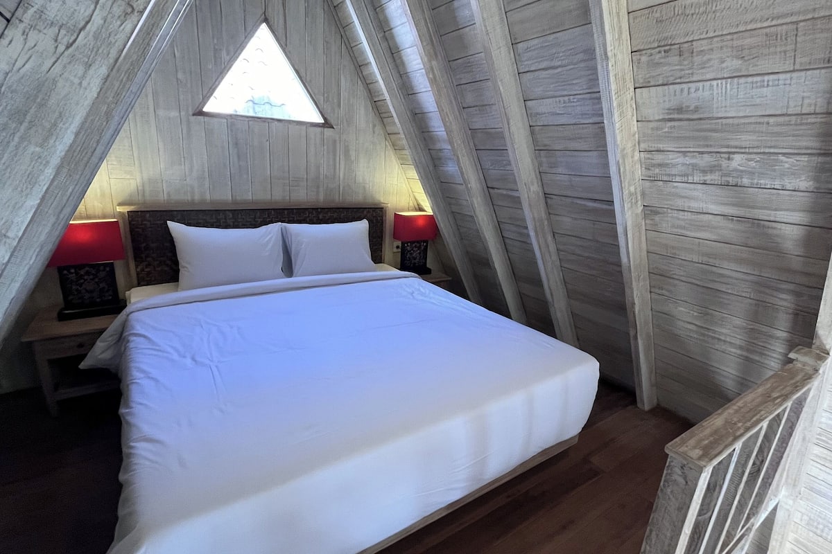 A bedroom with a wooden sloped ceiling, a bed with white bedding, two bedside tables with lamps, and a small triangular window.