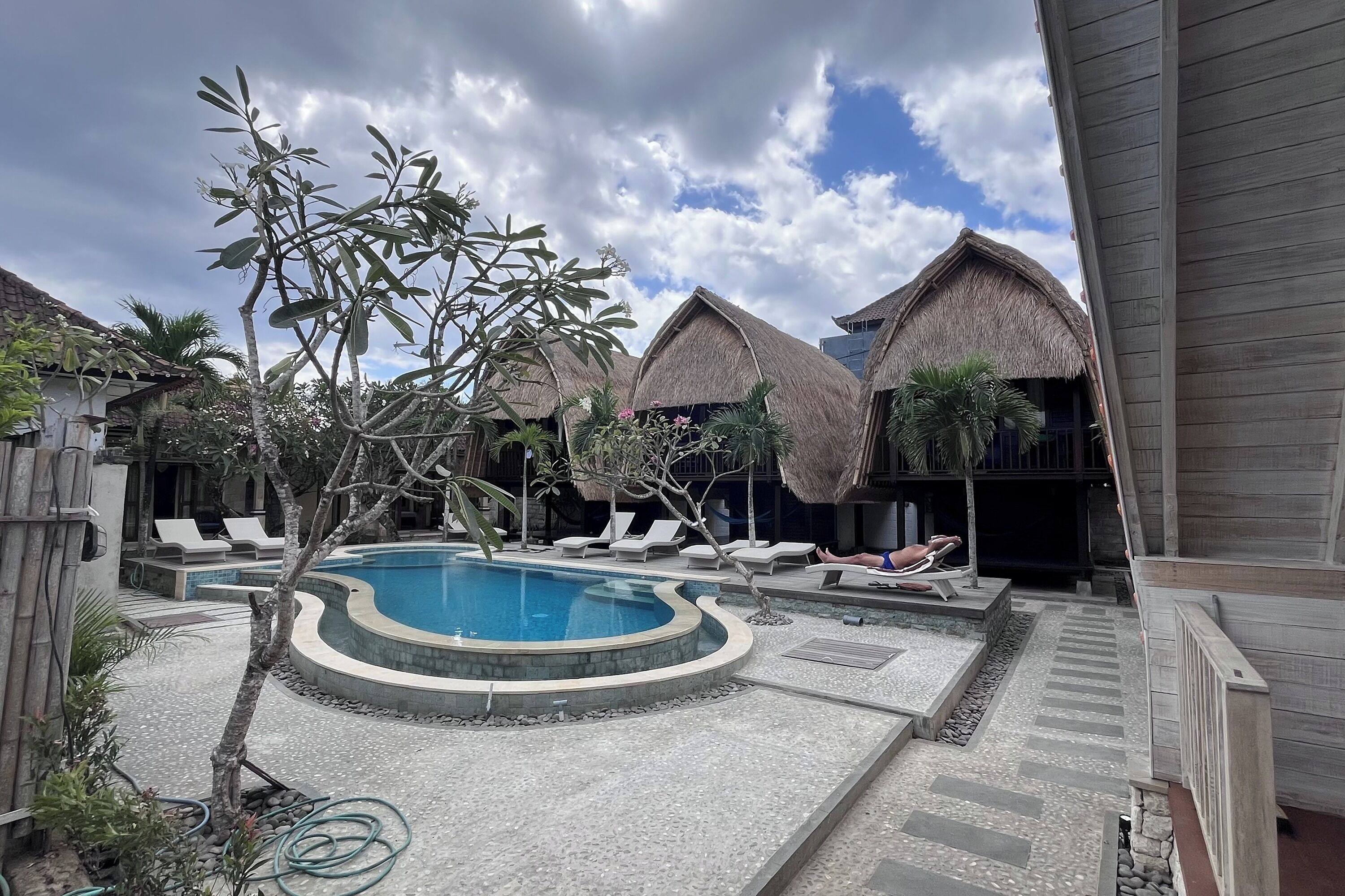 A pool area with lounge chairs, a thatched-roof building, and a small tree.