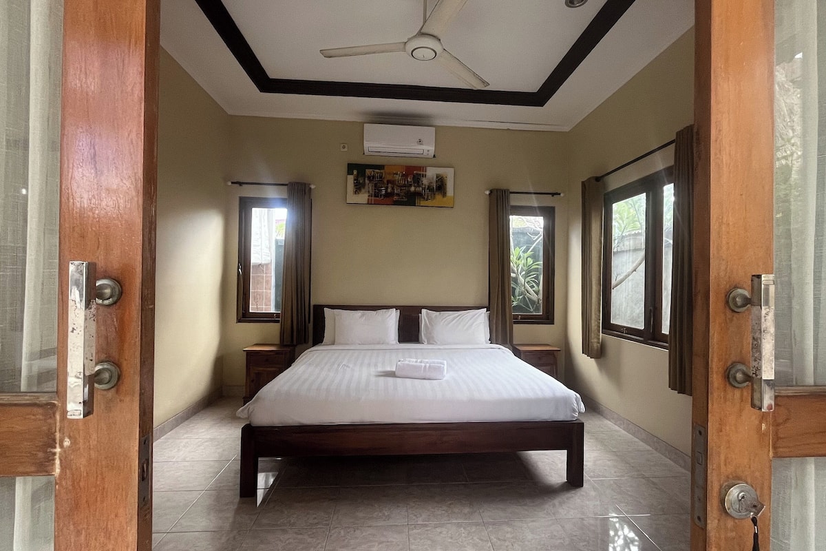 A hotel room with a large bed, wooden headboard, two bedside tables, an air conditioning unit, and large windows with curtains.
